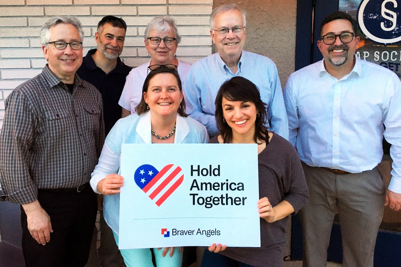 Seven Braver Angels members smiling outside a brick building, two in front holding a Hold America Together yard sign with the flag heart
