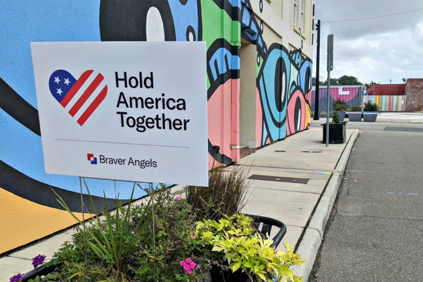 Hold America Together yard sign with the flag heart and Braver Angels logo, staked in a planter on a street beside a colorful mural