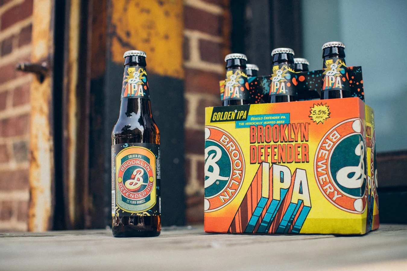 Brooklyn Defender IPA bottle and orange, yellow and blue six-pack carton on a stone ledge in front of a brick wall