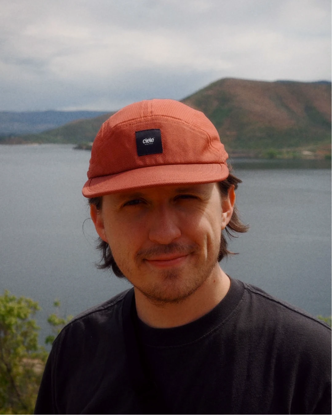 Sam Reed in a rust cap and black T-shirt above a lake and green hills