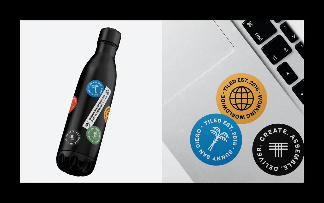 Black water bottle covered in round stickers beside a laptop with gold, blue and black Tiled stickers, on white